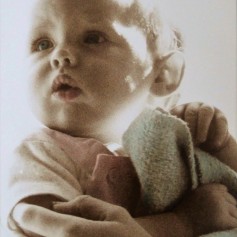 A delicate photo of a baby being held and looking to one side. The photo has been chemically sepia-toned. The baby's eyes and blanket have been hand-tinted blue, her mouth and clothing pale pink.