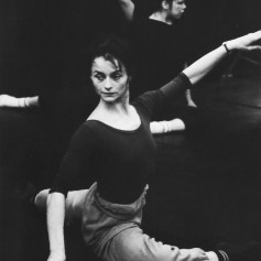 Black and white photo of a female ballet dancer in rehearsal. She's sat on floor in a ballet position with one arm outstretched. There is another dancer just out of focus behind.