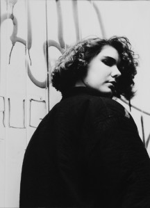 Black and white photo of a teenage girl looking back towards the camera, over her shoulder. Her pose and expression convey attitude. She's wearing a dark jacket and has shoulder length curly hair. The background is a wooden door covered in graffiti.