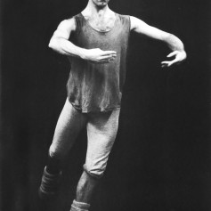 Black and white photo of a male dancer against a dark background. He is wearing a loose shirt, leggings and a bandana. A focused expression on his face as he holds a simple ballet position.