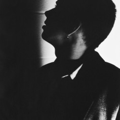 Stylish head and shoulders black and white shot of a guy with a flat-top hair cut. Photo is sparsely lit. Subject is almost silhouetted and is looking towards left of photo which is in total shadow.