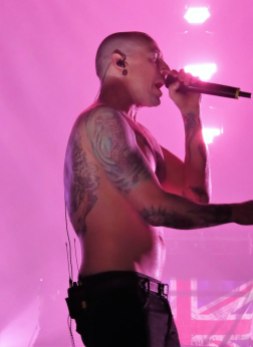 Side profile of Chester on stage, waist up, shirtless, showing his tattoos. He's singing against a backdrop of pink stage lighting.