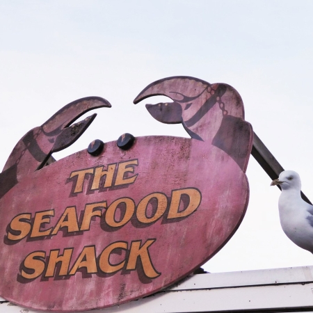 A pink crab-shaped sign with orange writing saying 'The Seafood Shack'. Next to it are two gulls poised for oppportunity.