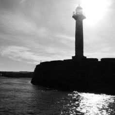 A dramatic black and white view of the sea and a lighthouse. The sun is glaring brightly, looking as though it is being emitted by the lighthouse.