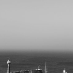 Black and white photo looking out to sea from a high viewpoint. Two piers with lighthouses fill the lower foreground. The sea and mist merge giving an eerie greyscale effect.