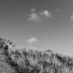 Black and white photo of a grassy hill with a dirt path leading to a pillbox fortification. Shot against a sky with a few clouds.