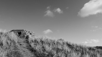Black and white photo of a grassy hill with a dirt path leading to a pillbox fortification. Shot against a sky with a few clouds.