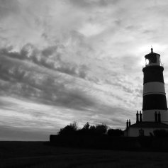 Black and white landscape photo of a striped lighthouse almost silhouetted against a dramatic cloudy sky.