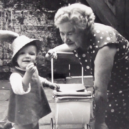 Black and white photo of me as a toddler with my Grandma. We are both kneeling in a backyard, with a toy pram between us. I'm wearing a dress and straw hat. My Grandma is in a spotty dress and smiling at a sweet I'm holding.