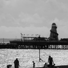 Black and white photo of a stone beach looking towards the sea and the end of a pier. There are silhouetted figures on the beach engaged in a variety of seaside activities. There is a helter-skelter and flags on the pier, set against a cloudy sky.