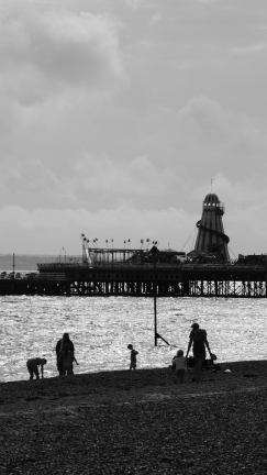 Black and white photo of a stone beach looking towards the sea and the end of a pier. There are silhouetted figures on the beach engaged in a variety of seaside activities. There is a helter-skelter and flags on the pier, set against a cloudy sky.