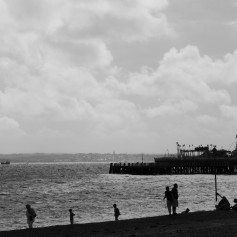 Black and white photo of a wide silhouetted beach scene. There are figures on the beach and a helter-skelter on a pier which is jutting out to sea. In the hazy distance are the hills of an island, all under a cloudy sky.