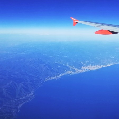 View through plane window looking down on Genoa and the edge of The Alps. The wingtip is visible against a hazy blue sky. Deep blue sea takes up the bottom right corner.