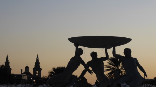 Silhouetted fountain against a pale sunset sky. The fountain comprises three Tritons (Mermen), holding aloft a large basin. To the left is the silhouette of a church and its bell towers.