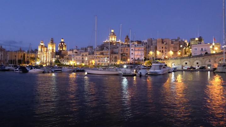 View from ferry back to harbour and marina at dusk. Yachts in the foreground against a backdrop of lit buildings and churches. Their lights are reflected in the water.